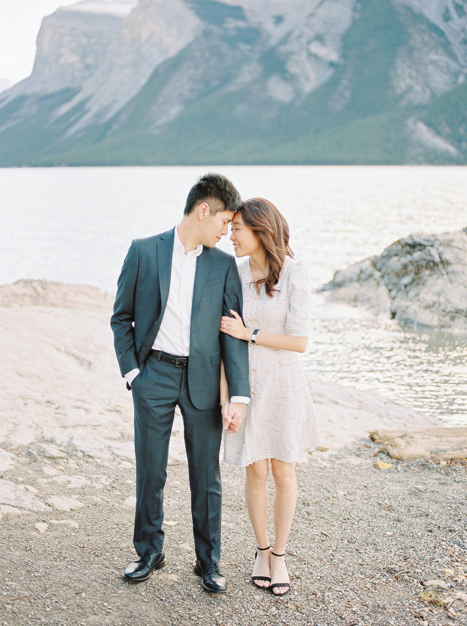 a classy engaged couple embrace while the sun rises over Banff Lake Minnewanka