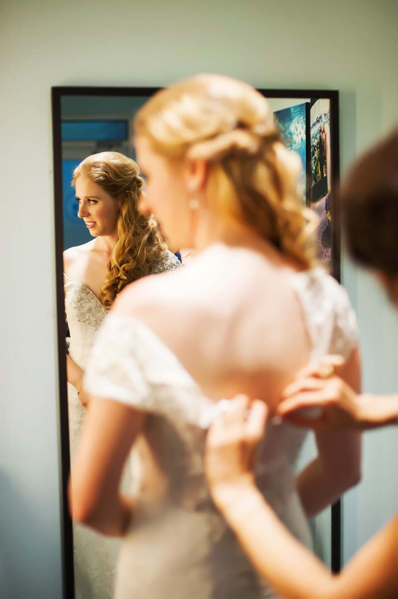 woman buttoning bride's wedding dress in front of mirror