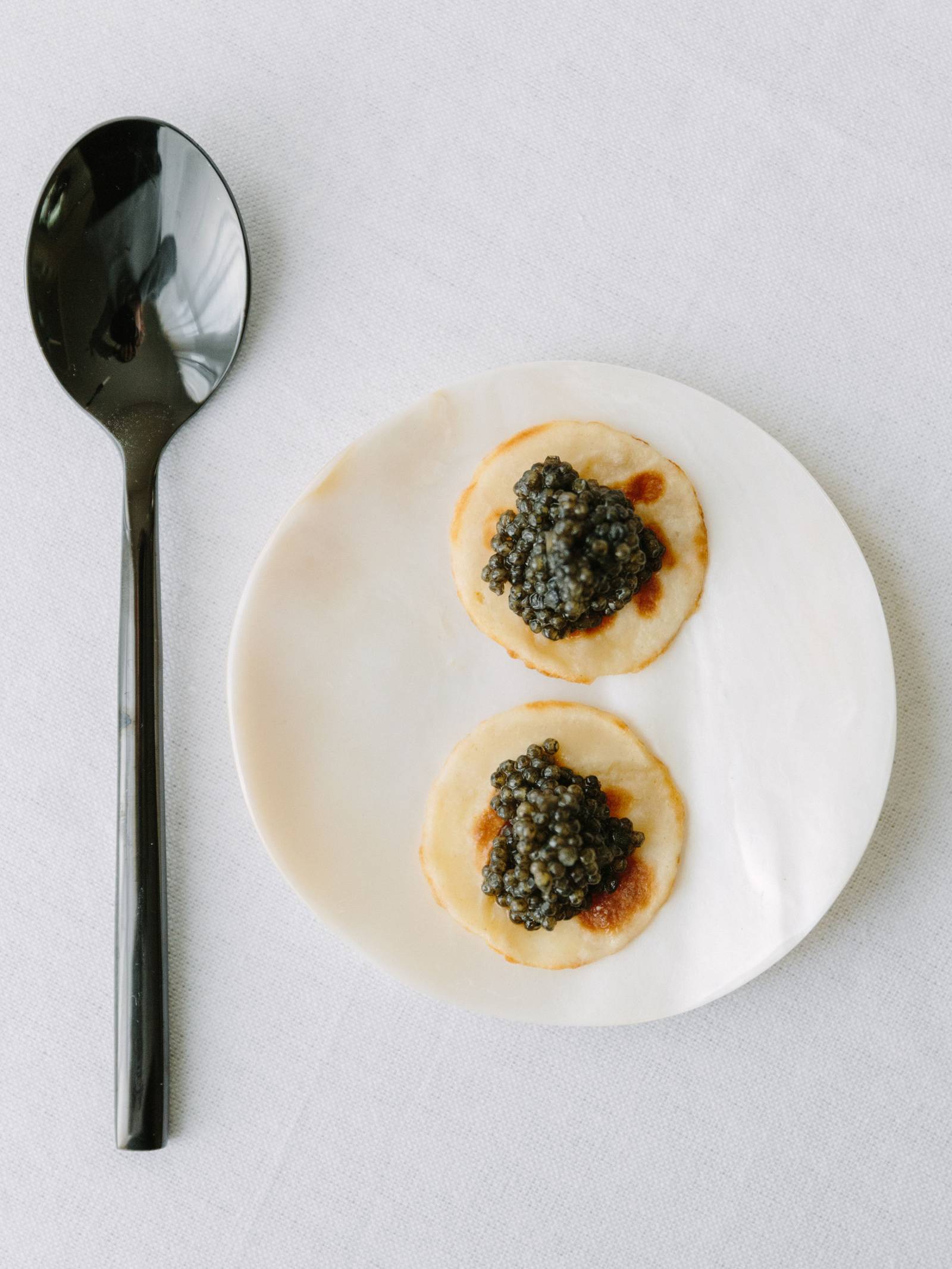 Black spoon next to white plate with caviar on blinis.