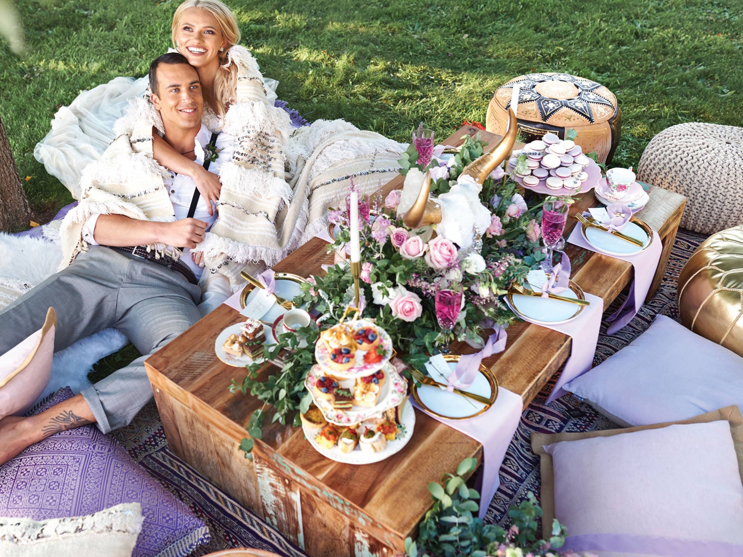 Bride and groom lounging by rustic wood table with pink and lavender florals, desserts and china