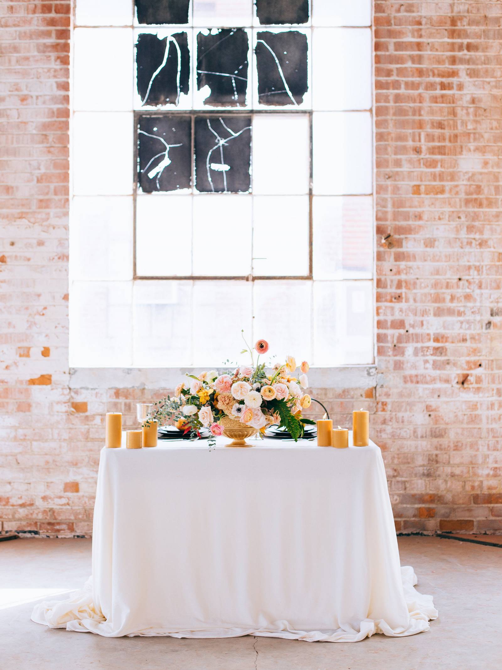 Sweetheart table with centerpiece of pink, yellow and coral in front of warehouse windows