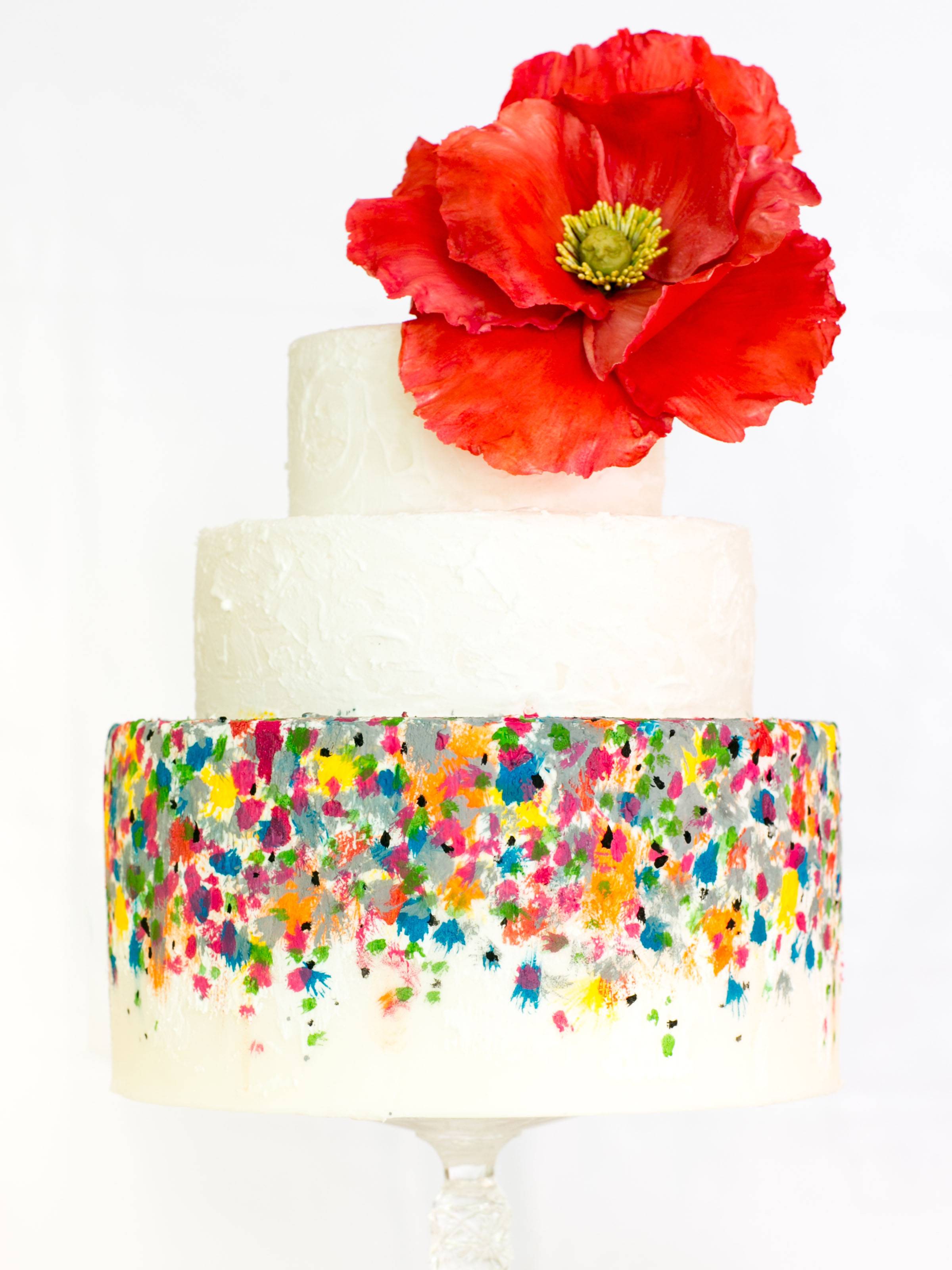 round, three-tiered wedding cake with confetti icing and large red poppy cake topper