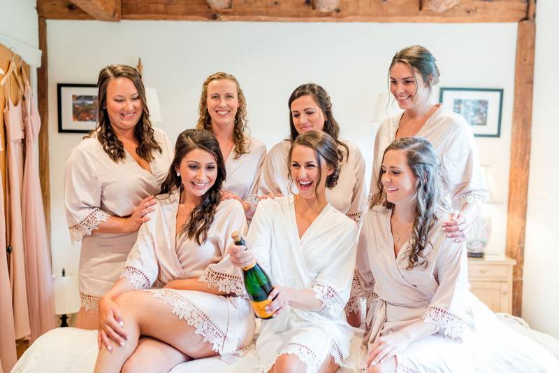 Bride and bridesmaid sitting on bed and popping champagne