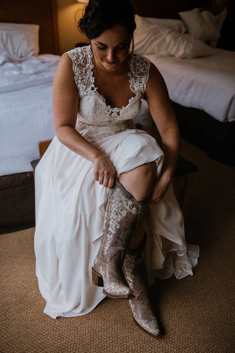 Bride wearing lace dress putting on wedding cowboy boots for rustic barn Vermont wedding