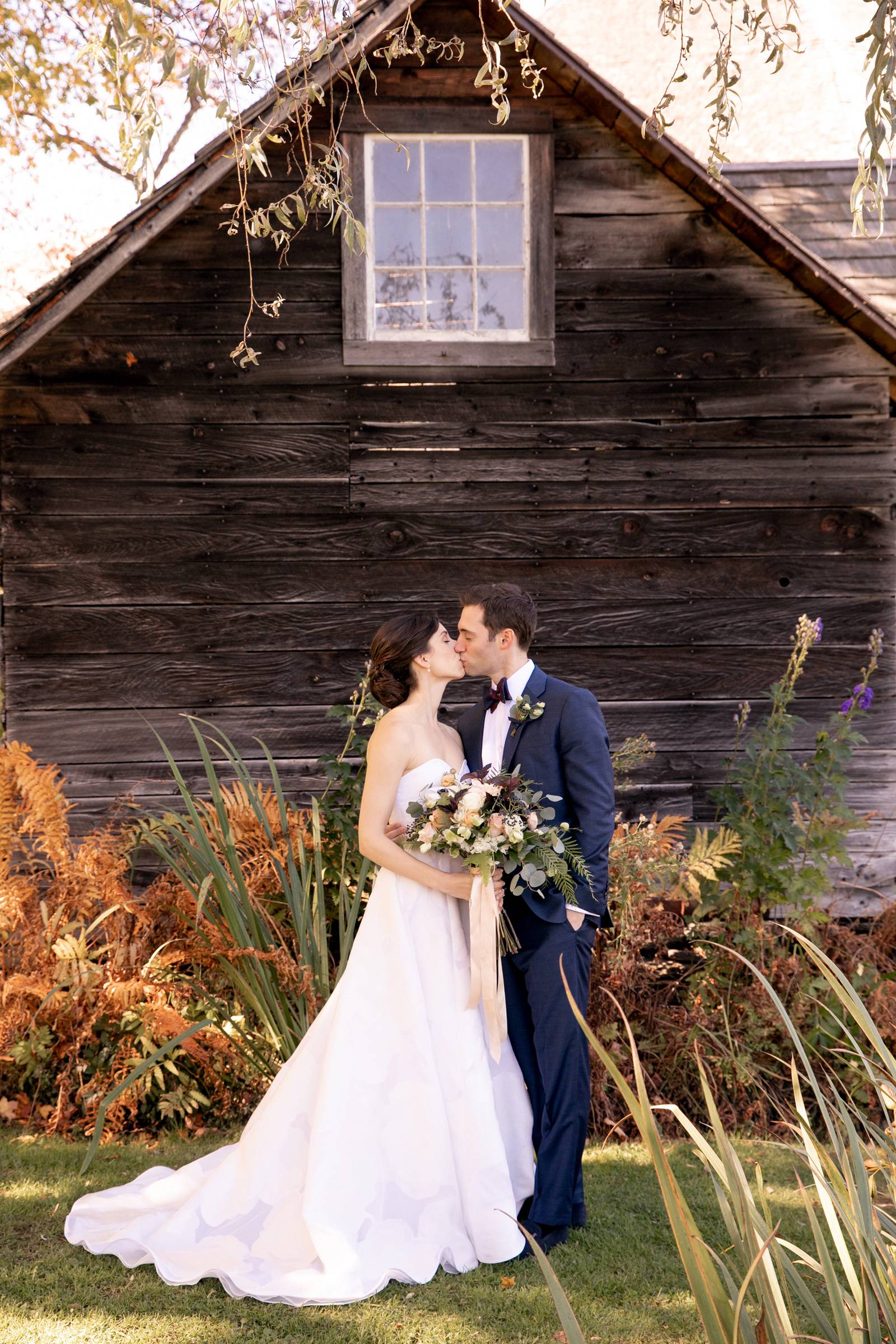Classic bride and groom kissing portrait in front of rustic barn on Vermont wedding day at the Inn a