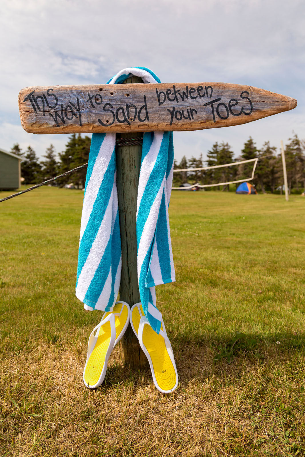 Wooden sign that says This Way to Sand Between Your Toes with a blue and white striped towel and yel