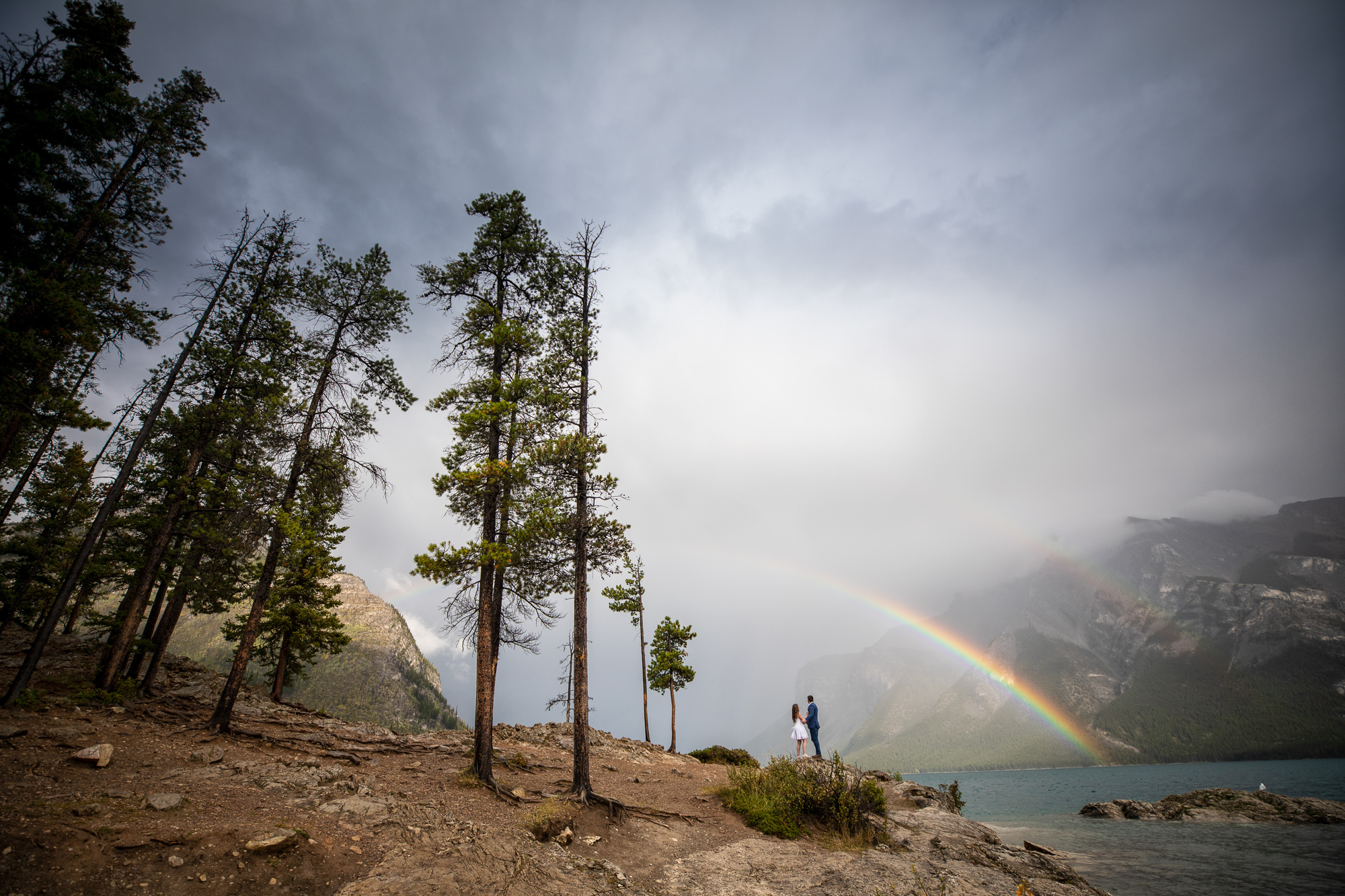 Couple sharing a romantic kiss under a vibrant rainbow at Minnewanka Lake in Banff, surrounded by th
